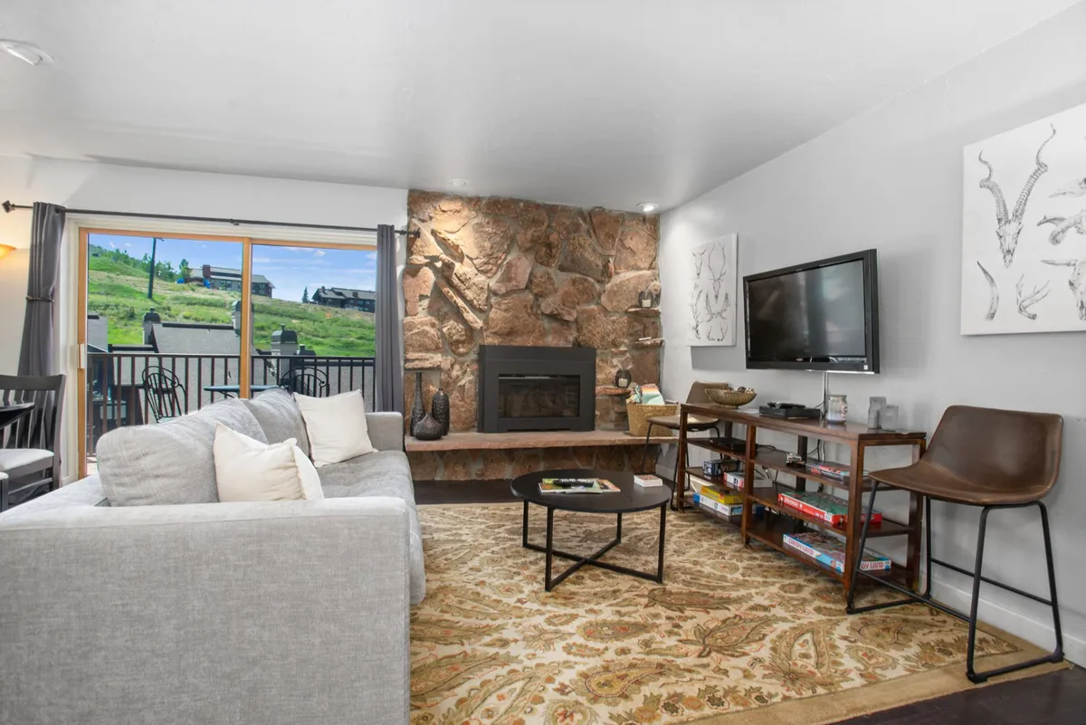 An inviting interior living space with a stone fireplace, plush sofa, and a large window looking out over green hills (or, seasonally, snow-covered slopes). The room is bright and welcoming, ideal for warming up after skiing, enjoying modern comforts, and watching the sun set over the mountains.
