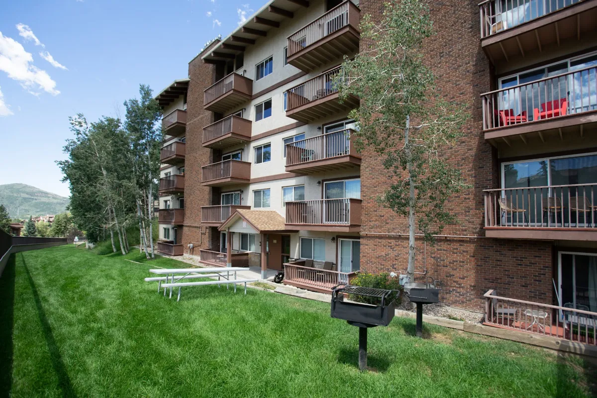 This exterior shot shows the apartment building with a green lawn, barbecue grills, and balconies. The image features people relaxing on balconies, children playing in the yard, and a sense of community with outdoor cooking and social gatherings taking place.
