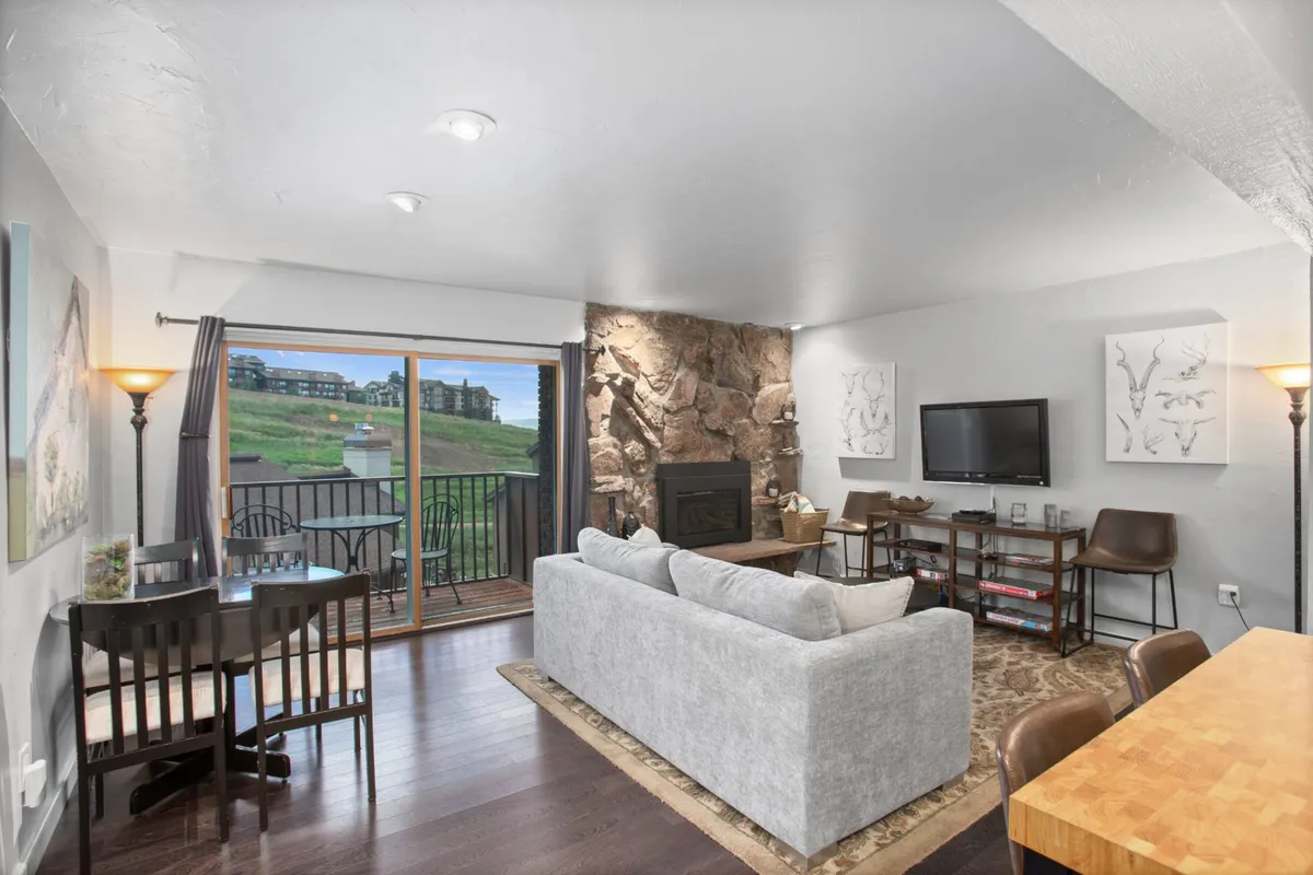 A spacious open-plan living and dining area featuring a comfortable sofa, stone fireplace, large window with mountain views, and a dining table. The arrangement encourages both relaxation and social interaction, and the window draws attention to the beautiful scenery outside.