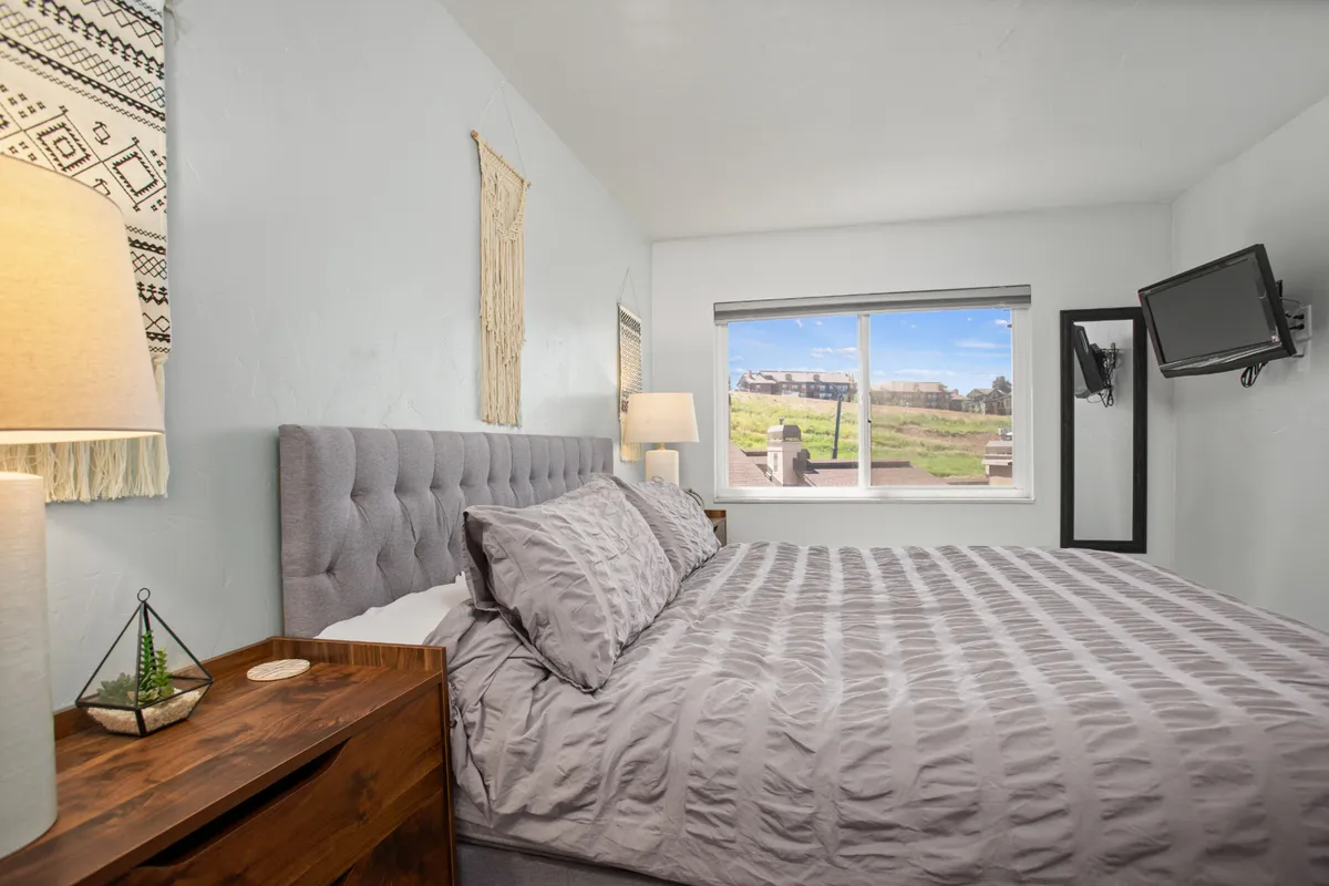 This interior scene showcases a tranquil bedroom with a plush bed, soft lamp lighting, a window allowing in natural light, and a television across from the bed. The setup exudes comfort and modern convenience.