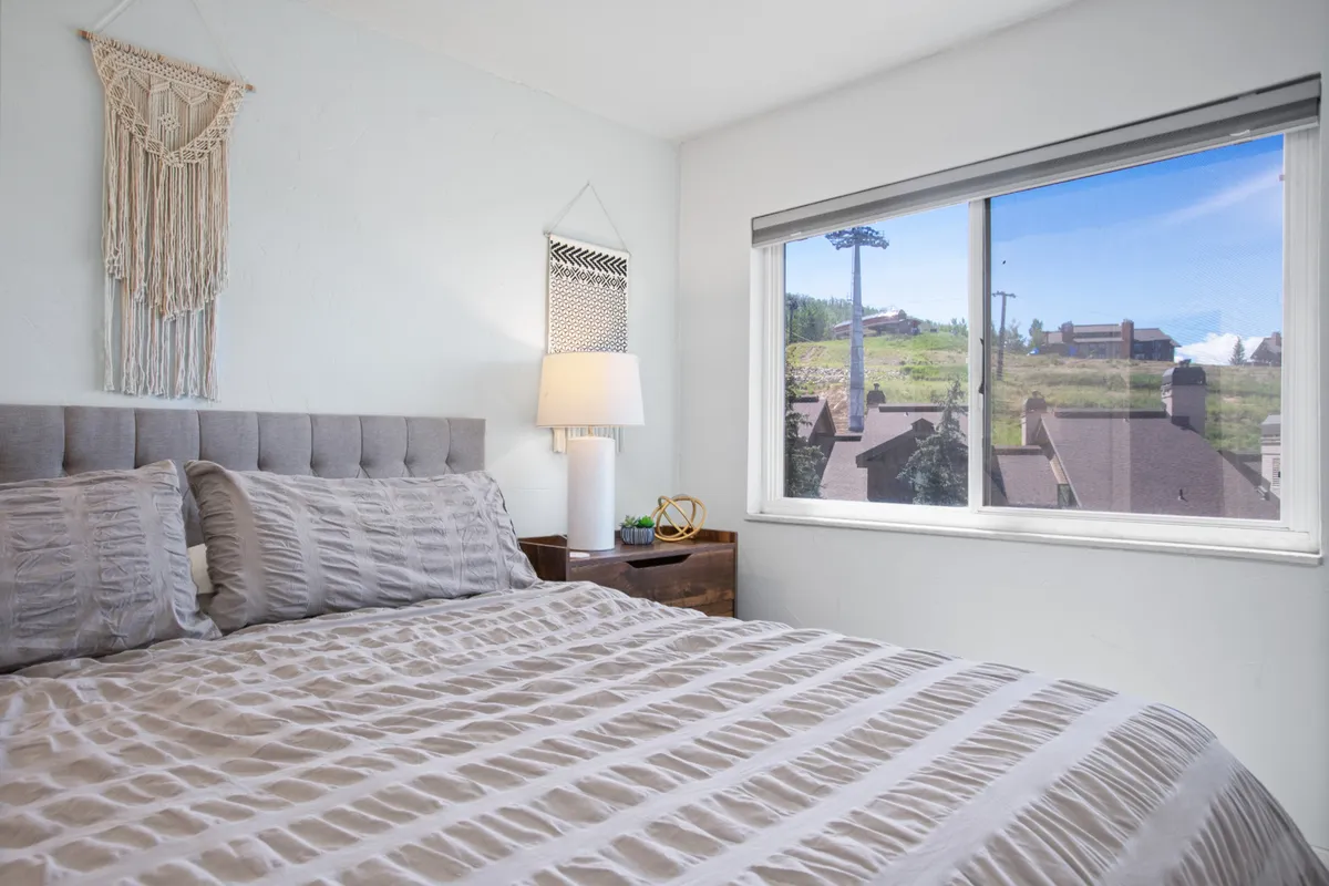A soothing, inviting bedroom featuring a bed with textured bedding, a window with a scenic view, a decorative wall hanging, and a nightstand with a soft-glowing lamp. The overall feel is calming and elegant.