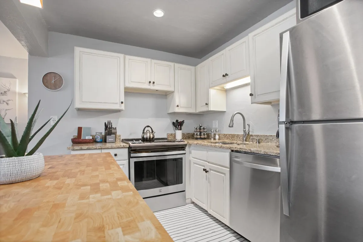 A close-up of the kitchen workspace featuring a granite countertop, shiny stainless steel appliances, white cabinetry, a wooden cutting board, and a decorative plant. The counters are free of clutter, inviting guests to cook, bake, or prepare snacks with ease.