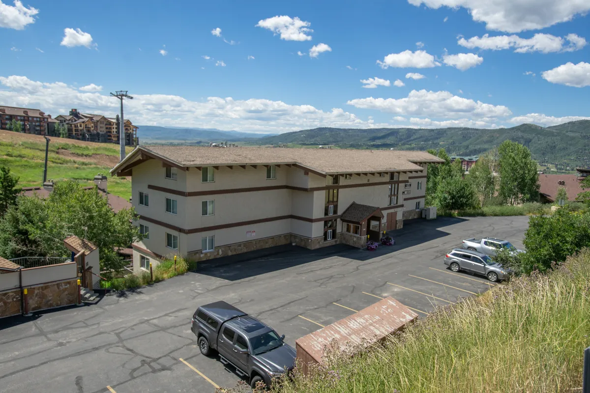 This image captures the exterior of the building against a scenic mountain backdrop, with visible greenery and vehicles in the parking lot. The landscape surrounding the structure hints at immediate trail access and a welcoming environment for outdoor activities like biking and hiking.