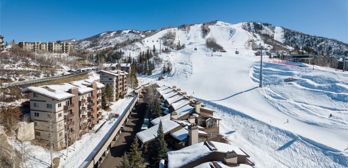 A sweeping exterior view of the condo complex, set against snow-covered slopes, with nearby ski lifts and a dramatic mountain backdrop. Residential buildings are visible, nestled among wintry trees, evoking a sense of both adventure and cozy retreat.