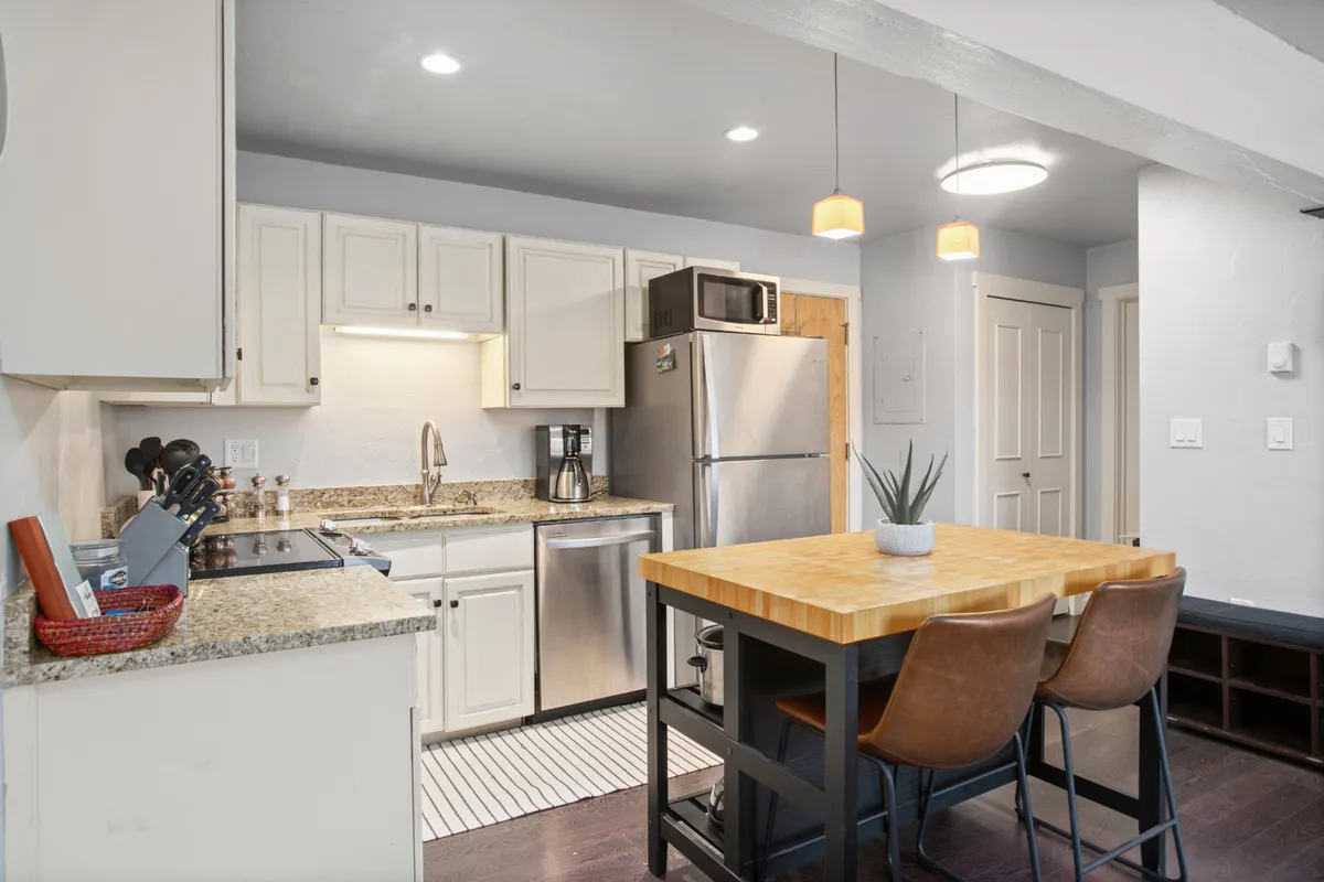 A view of a stylish kitchen and dining area showcasing granite countertops, a wooden table set for four, bar stools, and modern stainless steel appliances. The space is open and encourages interaction, ideal for both preparing and enjoying meals together.