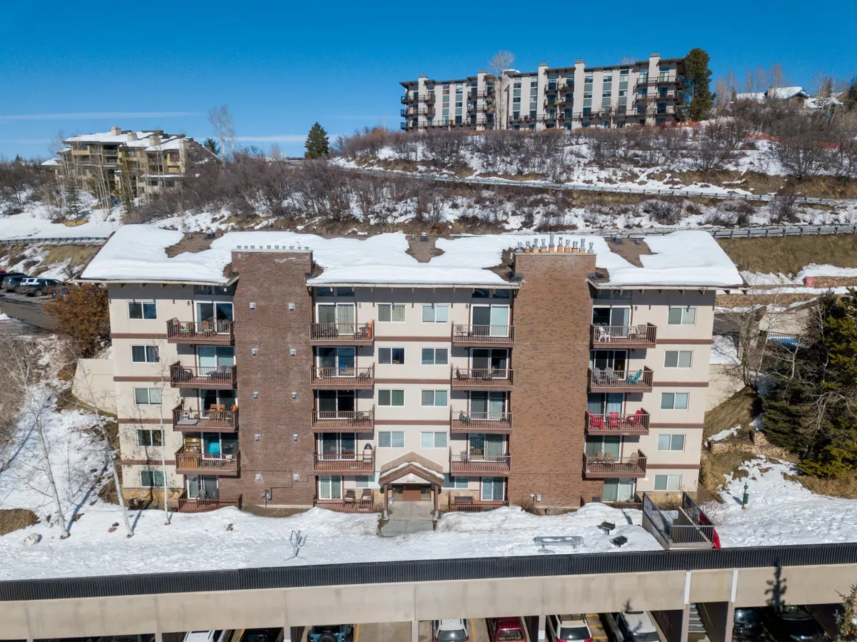 This exterior shows the apartment building in a winter setting, with a snow-covered roof and surrounding landscape. The parking area is visible, suggesting accessibility, and the setting evokes thoughts of ski trips, holiday gatherings, and cozy returns after outdoor activities.