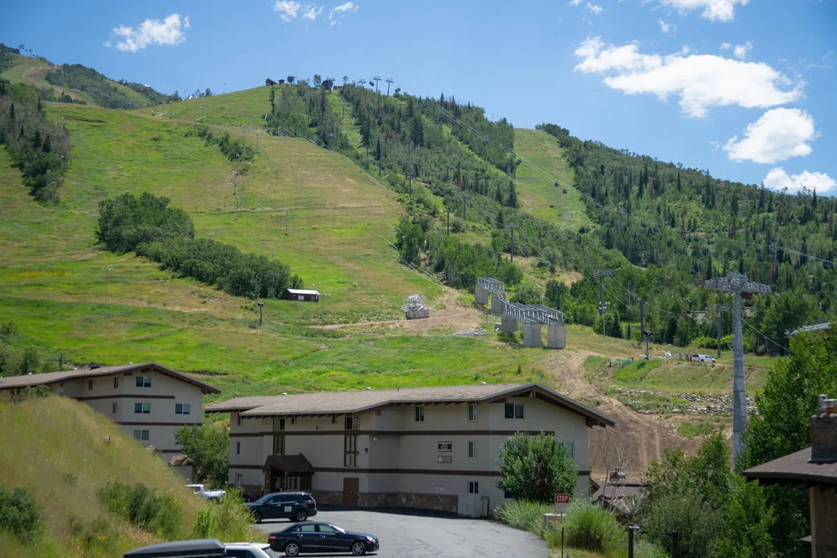 A wide view of the mountain range with open green spaces, buildings, and a ski lift. The lush landscape during summer invites visitors to hike, picnic, and experience nature up close, with trails leading into the distance and the resort nestled at the base.
