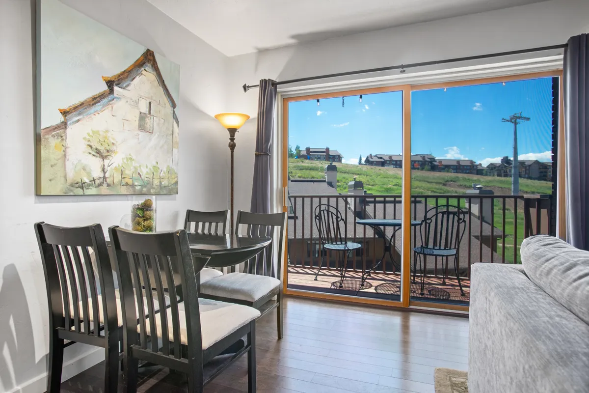 A welcoming dining area with a wooden table and chairs, artwork on the wall, and a view onto a balcony with mountains beyond. The image captures the intimate atmosphere suitable for shared meals and conversation, with natural light enhancing the scene.