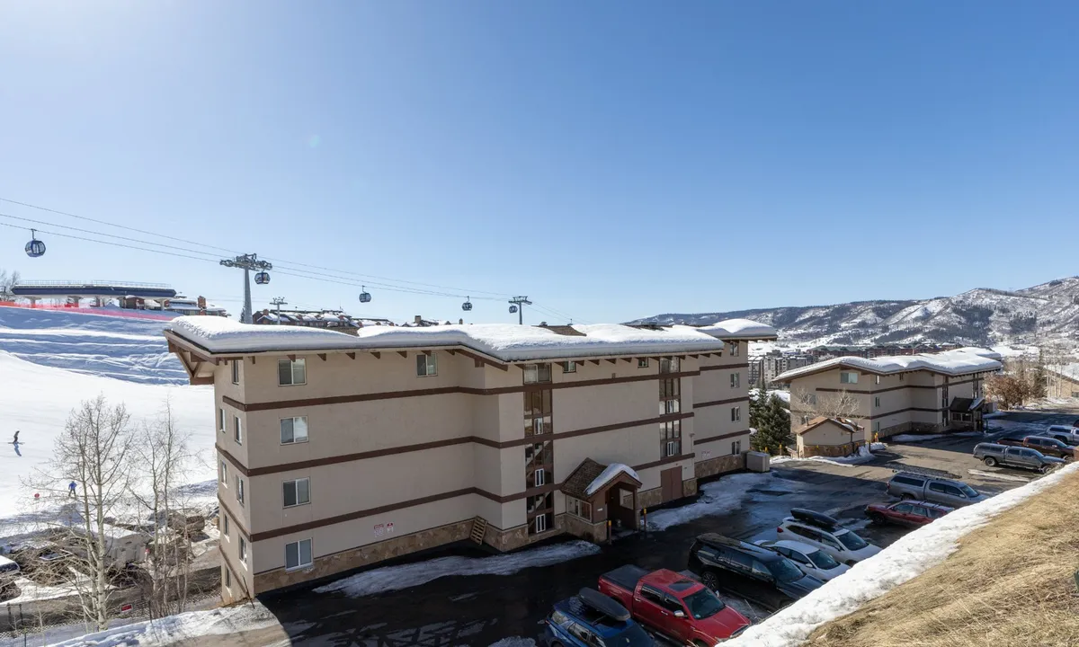 This image captures the essence of ski-in/ski-out living with visible ski lifts, snow-covered slopes, apartment buildings, and vehicles in the parking lot at the base of the mountain. The atmosphere is lively and adventurous, showcasing immediate access to the slopes and vibrant après-ski gatherings at the foot of the hill.