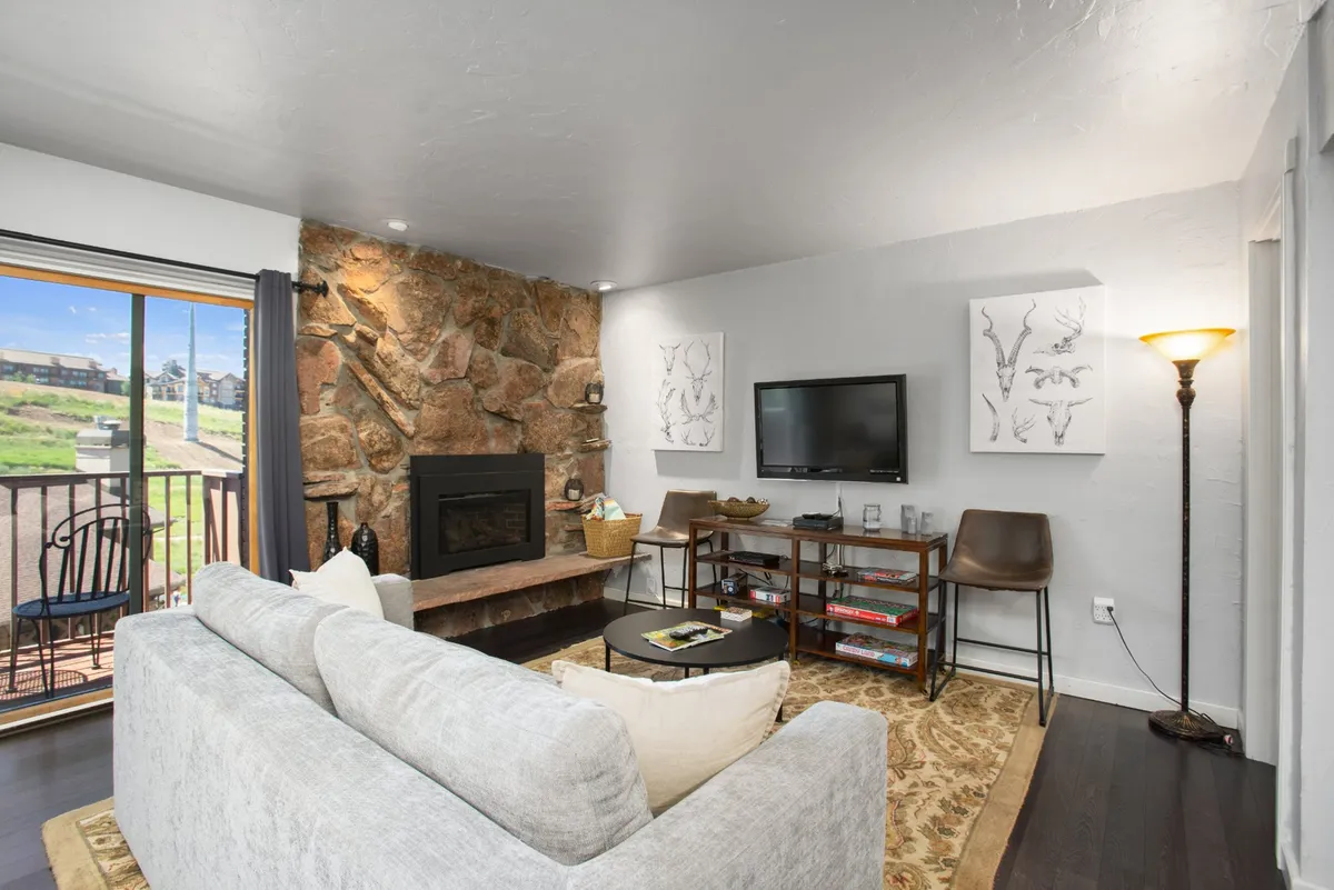 The image depicts a warm and inviting living area inside the condo, featuring a beautiful stone fireplace as the centerpiece. Large windows flood the room with natural light and offer a scenic view, possibly of the Colorado mountains. Thoughtful artwork adorns the walls, and a comfortable coffee table setup invites guests to unwind. The overall mood is cozy and welcoming, perfect for relaxing after outdoor adventures.