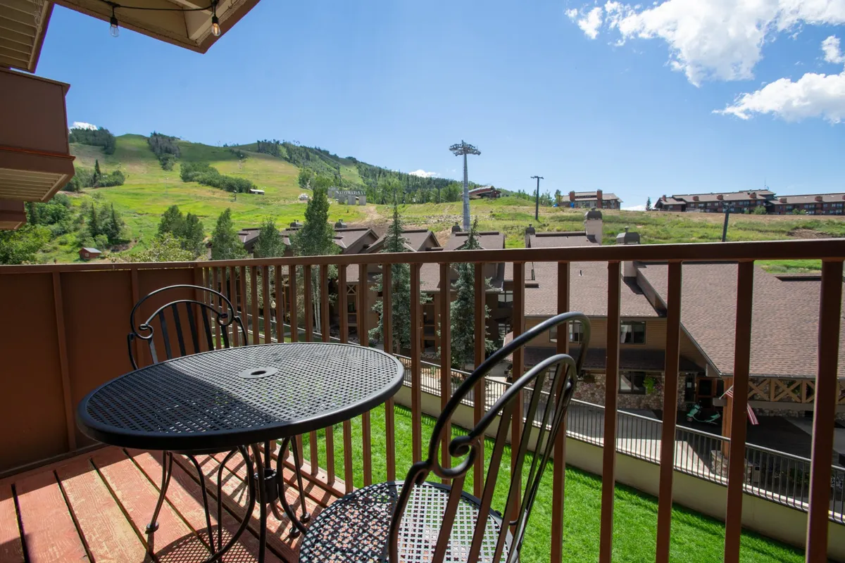 A tranquil balcony scene featuring a small table and chairs, perfect for two, overlooking a breathtaking mountain view with nearby buildings softly receding in the background. The soft lighting hints at either early morning or late evening, enhancing the sense of peaceful seclusion.