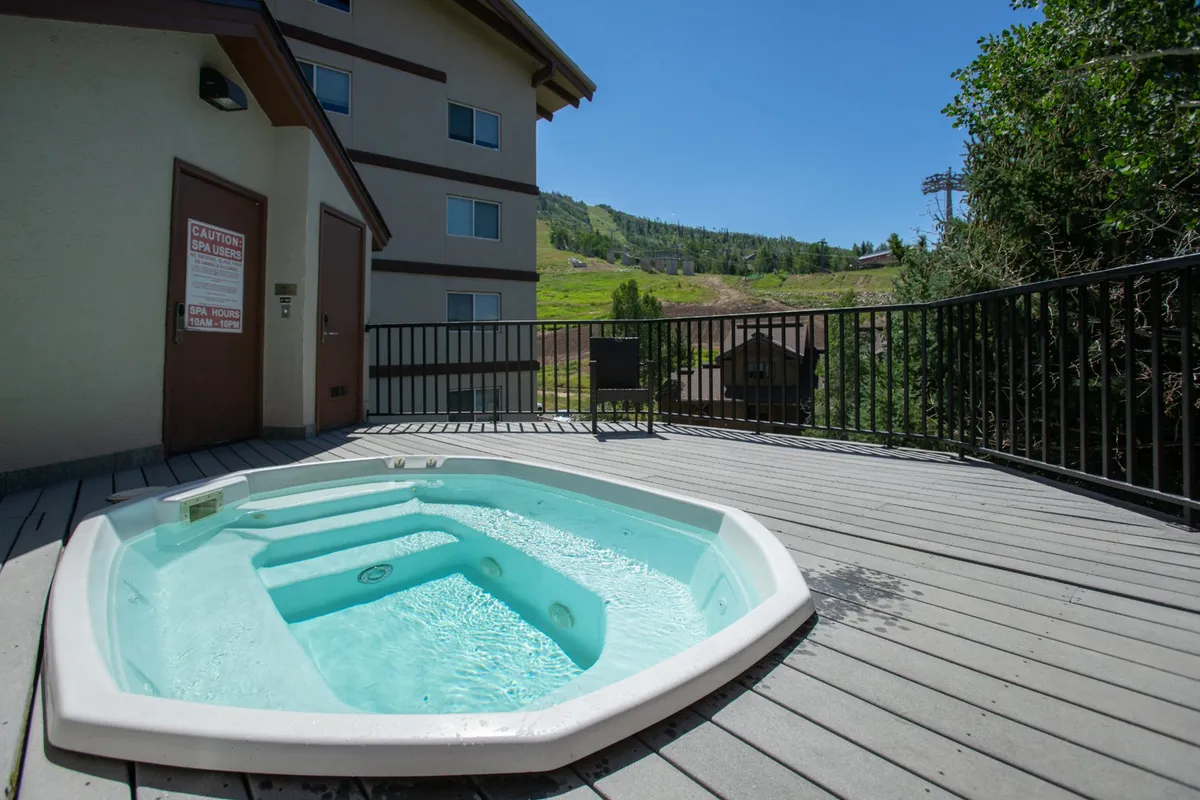 An inviting outdoor hot tub sits on a spacious deck with sweeping views of the mountains. The setting is tranquil, perfect for relaxing after a day of adventure. A few guests are seen enjoying a soak, framed by the glow of the sunset and the natural beauty of the surrounding landscape.